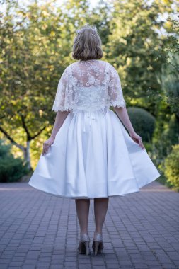 Bir gelinin, çay uzunluğundaki gelinliğinin kenarlarını tutarak pelerininin hassas dantel detaylarını ortaya çıkarması. Yeşilliklerle çevrili bir kaldırım taşının yolunda yakalandılar.