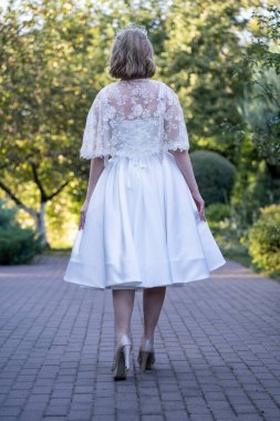 Bir gelinin, çay uzunluğundaki gelinliğinin kenarlarını tutarak pelerininin hassas dantel detaylarını ortaya çıkarması. Yeşilliklerle çevrili bir kaldırım taşının yolunda yakalandılar.