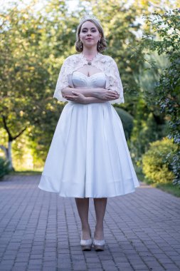 Beyaz çay uzunluğunda bir gelinlik, dantel bir pelerin ve taçla kaldırım taşından bir patikada duran bir gelinin tam boy portresi. Bahçe dekoruyla çevrelenmiş bu görüntü zarafeti yakalar.