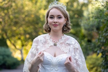 Beyaz dantelli gelinlik ve taç giymiş zarif bir gelin. Nazikçe dantel pelerininin kenarlarını tutuyor. Yumuşak doğal ışıkta bulanık arka planda, portre zamansız güzelliği çağrıştırıyor.