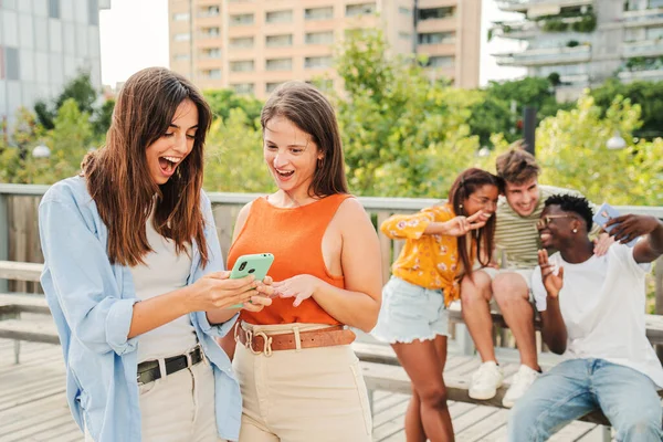 2人の魅力的で明るい笑顔の女性が大学キャンパスの外のスマートフォンで最後に面白い面白いと面白いニュースを見て示しています バックグラウンドでは 3人の若い友人 楽しみを持っている — ストック写真