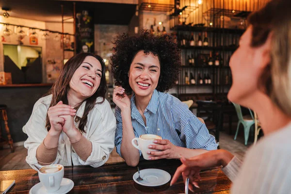 A group of young women talking and laughing sitting on a coffee shop or restaurant. Two ladies smiling and looking his friend holding a cup. Three girls having fun cahtting on a bar. Lifestyle concept