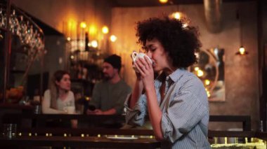 Young hispanic woman with curly hair drinking coffee in a restaurant, in the background a group of friends talking. Latin happy girl smiling and looking ant camera with a coffee cup enjoying the relax