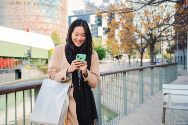 Young beautiful asian woman smiling typing on smartphone app a message. Portrait of one happy chinese girl using a cellphone after shopping on a city street. Connection concept. High quality photo
