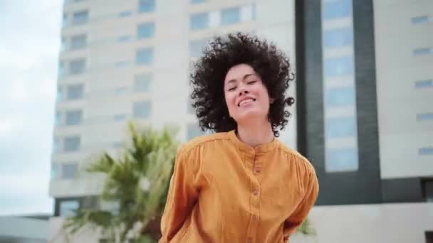 巻き毛とオレンジのシャツを着たクレイジーヒスパニック系の若い女性が幸せを表現して屋外で音楽を踊っています ラテン系の可愛い娘が街中でダンスをしてる 自由の概念 高品質4K — ストック動画