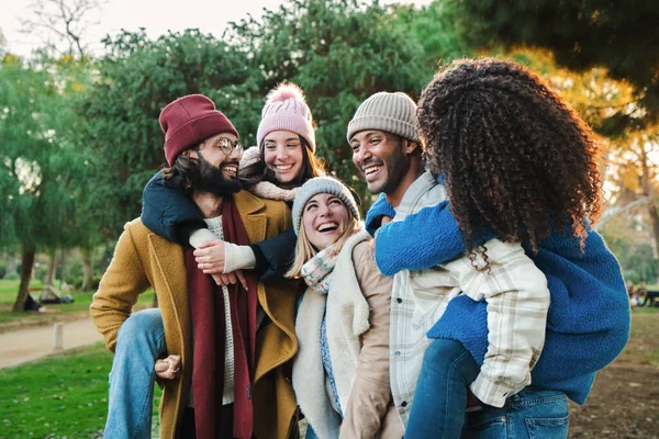 A group of happy young friends enjoying a weekend trip having fun together in a natural park. Five multiracial people smiling and having a affectionated piggybacks to their girlfriends. High quality