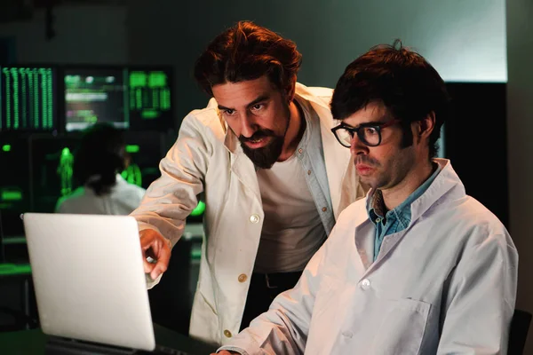 Two scientific male doctors with lab coat doing a medicine, biology or pharmacy investigation and looking concentrated speaking aboutto the laptop screen. Work team of scientist talking about medicine