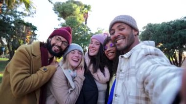 Group of young friends waving and having fun taking a selfie portrait or video call with a smartphone in a park, smiling happy people laughing taking a photo looking at camera outdoors in autumnal