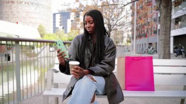 Gülümseyen mutlu Afro-Amerikalı kız dışarıda bankta cep telefonuyla alışveriş yapıyor. Örgülü ve hippi kıyafetli genç bir kadın. Şehir caddesinde akıllı bir telefonla mesajlaşıyor.