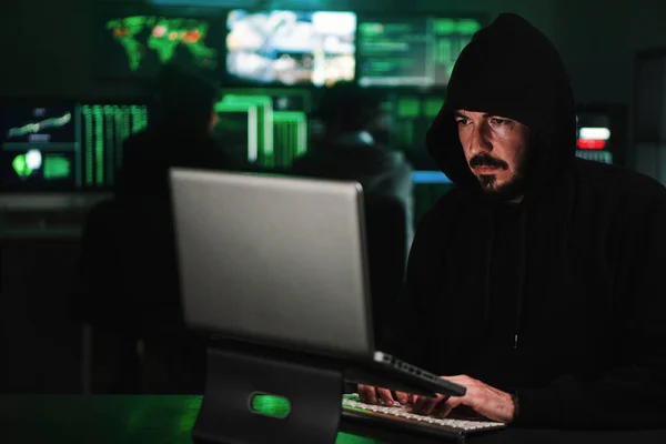 Hooded male criminal hacker using a laptop to breack the security of the firewall, at background a lot of computer screens. Pishing concept. High quality photo