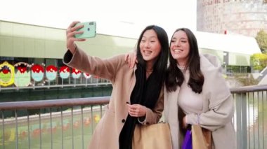 A couple of young women having fun and posing doing a selfie portrait with a cellphone, two girls taking a photo with a smart phone on a shopping day in the city mall. Lifestyle concept. Slow motion
