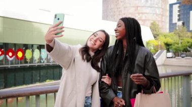 Two multiethnic young women smiling and having fun taking a selfie portrait with a smart phone after shopping and sharing in the social media. A couple of carefree girls doing a photo with a cellphone