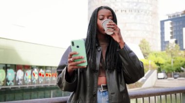 Young african american girl with braids watching videos on a social media using a smartphone drinking a espresso coffe standing outdoors. Communication concept. Slow motion. High quality 4k footage