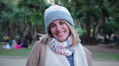 Front view of one happy caucasian blonde young woman smiling with white perfect teeth, wearing a beanie hat and looking excited to the camera in a park outdoors. Slow motion. High quality 4k footage