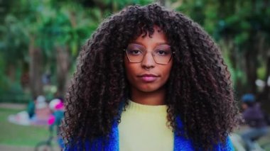 Close up portrait of young serious african american woman with goggles and curly hair looking pensive at camera with sad attitude. Front view of multiracial girl standing at park outdoors. Slow motion