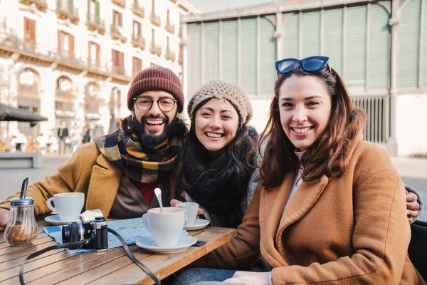 Group of happy friends taking a break drinking a coffee cup in a restaurant terrace, having fun and looking at camera outdoors. Three tourist sitting outside of the bar smiling. . High quality photo