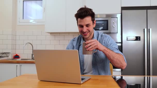 一个英俊的高加索人在家里的厨房里与笔记本电脑一起工作 一边喝咖啡 一边享受早晨的乐趣 快乐的年轻创业者或学生在网上搜索信息 自由应聘工作 — 图库视频影像