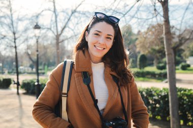 Portrait of young brunette woman smiling and looking at camera. Front view of a happy traveler girl with perfect white tooth standing outdoors with carefree attitude. Tourism concept. High quality