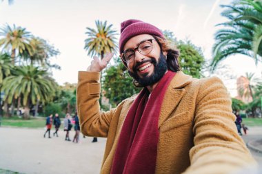 Bearded young caucasian male taking a selfie portrait with a smartphone in a park outdoors, wearing a beanie hat, glasses and scarf in autumn season. Close up photo of a happy man in a tourism trip