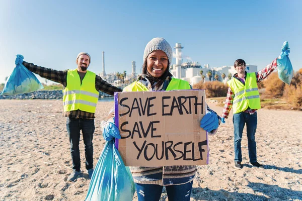 On foreground one happy environmental volunteer smiling and holding a poster cardboard with positive message, save the earth save yourself, at background her friends cleanup trash of the beatch sand