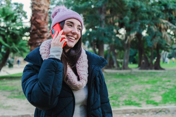 Young brunette woman with beanie hat calling smiling using a cellphone at park outdoors. Teenage girl talking using an mobile phone with autumnal clothes. Communication concept. High quality photo