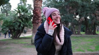 Açık hava parkında cep telefonuyla gülümseyen şapkalı esmer bir kadın. Sonbahar kıyafetleriyle cep telefonu kullanan genç bir kız. İletişim konsepti. Ağır çekim. Yüksek