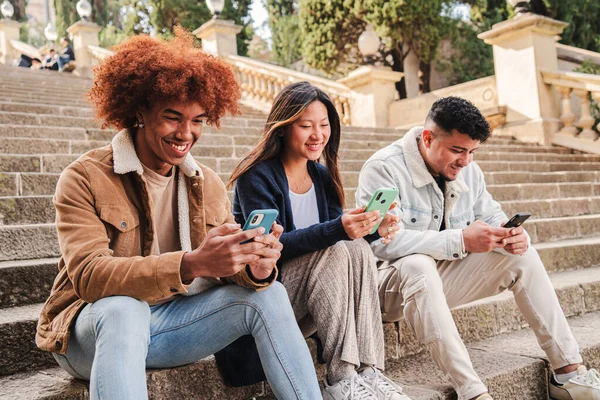 多人種グループの若い友人は 屋外の階段に座っている彼らの携帯電話アプリを使用して楽しんで笑顔 3つの幸せな多民族のティーンエイジャーは 彼らの携帯電話デバイスでゲームを楽しんでいます — ストック写真
