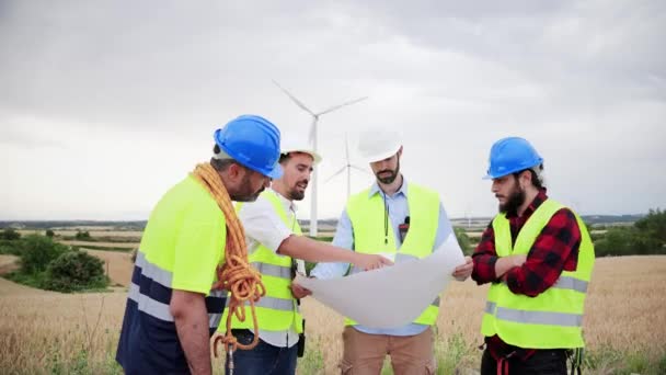 一组风车工程师正在观望涡轮机的计划 并在谈论涡轮机的建造或维修 穿着安全背心一起工作的专业技术人员 可再生能源概念 — 图库视频影像