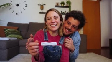 Mutlu beyaz bir çift kameraya pozitif hamilelik testlerini gösterdikleri için heyecanlı bir şekilde gülümsüyor. Karı koca, ellerinde bir kehanet tutarak bir bebek beklediklerini kutluyorlar.