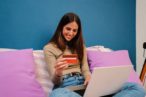 Beyaz tenli genç bir kadın dizüstü bilgisayar kullanarak oda ayırtıyor ve kredi kartıyla evin içindeki bir kanepede oturuyor. İnternetten bir kadın satın alımı. Finans konsepti. Yüksek