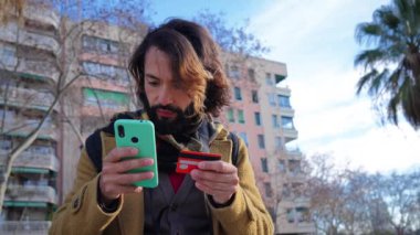 Genç bir adam kredi kartıyla alışveriş yapıyor ve dışarıda akıllı bir telefon cihazı tutuyor. Mutlu sakallı erkek teslimat için cep telefonu uygulaması kullanıyor ve finans bankacılığına bak. Yüksek