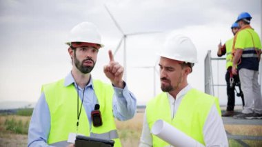 Bir grup rüzgar değirmeni mühendisi çalışıyor ve bir tablet kullanarak yenilenebilir enerji pervanesinin yapımını izliyorlar. Kask ve güvenlik yeleği giyiyorlar.