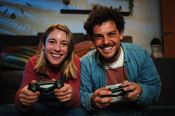 stock image Young couple having fun plaing video games with a console together sitting at couch at home. Two happy friends smiling and gaming at night holding the controller and touching gamepad buttons. High