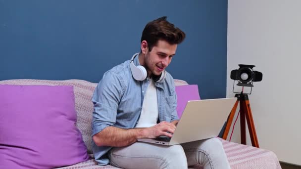 ソファの上に座ってカメラを見ているノートパソコンを持って家で働いているハンサムな起業家のフリーランスの男性 ワイヤレスインターネット接続のコンピュータを使用してインターネット上で閲覧若い男 — ストック動画