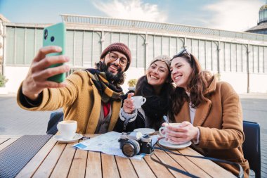 Bir grup arkadaş bir bar terasında ya da restoranda oturmuş dışarıda akıllı bir telefonla selfie çektirirken eğleniyorlar. Üç genç turist birlikte gülümsüyor ve cep telefonuyla fotoğraf çekiyor.