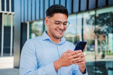 İspanyol genç girişimci ya da iş adamı gülümseyip cep telefonu uygulaması kullanıyor, yönetici gömleği ve gözlüğü takıyor. İş yerinde akıllı telefonuyla internette gezinen yakışıklı latin profesyonel erkek.