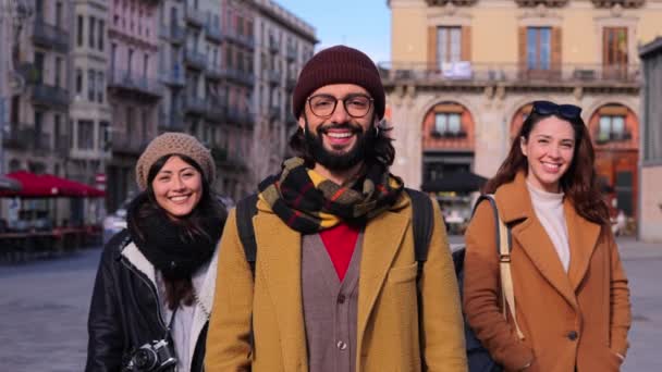 幸せな若者たちが一緒に笑みを浮かべて 屋外に立ってカメラを見てのグループの肖像画 ヨーロッパの街で旅行を楽しんでいる3人の有名なヒップスターの正面ビュー 手前に髭を生やした男が — ストック動画