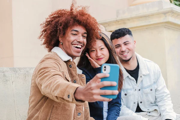 アフロヘアのアフリカ系アメリカ人の若者が 携帯電話のカメラを使って多人種の幸せな友人と自撮りの肖像画を撮るのを笑っていた スマートフォンアプリで写真を撮る人々のグループ — ストック写真