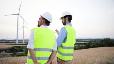 Bakım yapmaları gereken türbini gösteren iki rüzgar değirmeni mühendisinin arka görüntüsü. Yenilenebilir enerji teknisyenleri konuşuyor, çalışıyor, jeneratörü kontrol ediyor.
