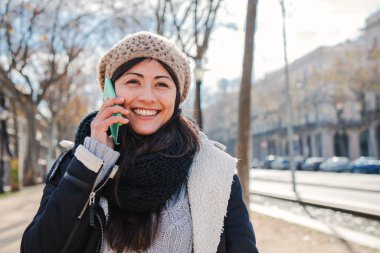 Dışarıda cep telefonuyla konuşan genç ve mutlu bir kadın. Gülümseyen ve cep telefonuyla konuşan Asyalı bir kadın. Şapka ve palto giyerek sohbet etmek için akıllı telefon kullanan neşeli bir bayan. iletişim