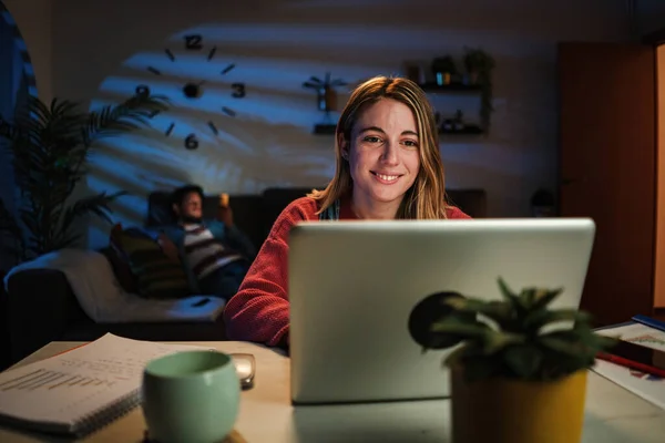 Young caucasian blonde woman working on her startup with laptop at home office sitting on desk at night. Happy female entrepreneur using a computer to browse on internet with wireless connection. High