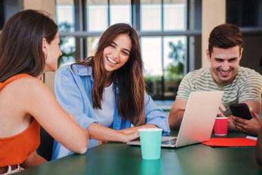 Bir grup gülümseyen genç öğrenci araştırma için dizüstü bilgisayar kullanıyor ve lise kütüphanesinde ödevlerini birlikte yapıyorlar. Üç mutlu Kafkas öğrenci arkadaş shool sınıfında oturmuş bilgisayarla öğreniyorlar.