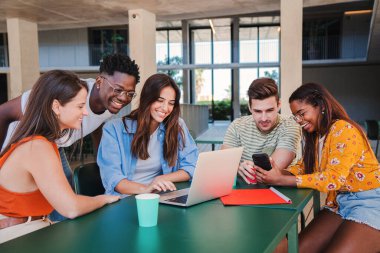 Bir grup gülen uluslararası öğrenci kütüphanede dizüstü bilgisayarla öğreniyor. Üniversite sınıfındaki ev ödevleri hakkında bilgi toplamak için bilgisayar kullanan gençler.