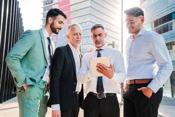 Bir grup yakışıklı iş adamı iş yeri binalarında bir tablet cihaz kullanarak takım elbiseyle çalışıyor. Başarılı bir avukat meslektaşlarına ekranı gösteriyor. Yönetici insanlar. Ortaklık