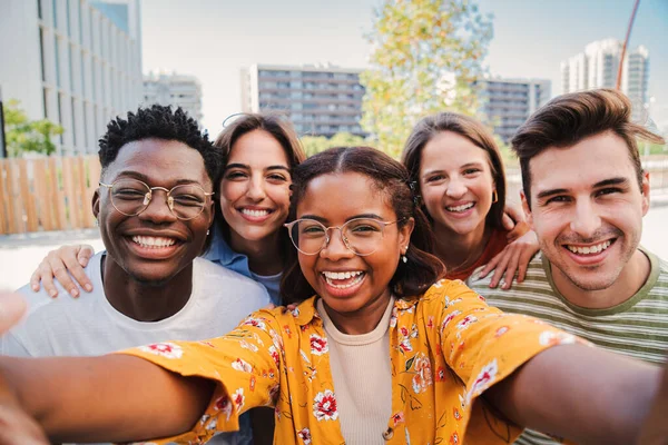 Üniversite kampüsündeki kameraya bakarak selfie çeken bir grup çok ırklı genç öğrenci. Mutlu Afro-Amerikan genç bir kadının neşeli arkadaşlarıyla gülüşünün yakın portresi.