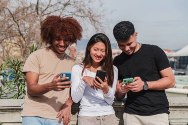 Bir grup çok ırklı genç arkadaş dışarıda cep telefonu aygıtı kullanarak eğleniyor. Gülümseyen ve gülen üç genç sokakta sosyal medyada cep telefonu paylaşımını seyrediyor.
