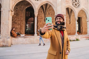 Gözlüklü, sakallı, gülümseyen ve sosyal medyada paylaşmak için bir Avrupa şehrinde selfie çektiren mutlu yetişkin bir adam. Kafkas turist erkek joutney gezisinde fotoğraf çekerken eğleniyor.