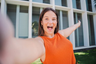 Selfie çekerken gülümseyen ve kollarını açan mutlu beyaz bir kadının yakın plan portresi. Güzel genç bir kadın. Üniversiteli neşeli bir öğrenci kampüsün dışında gülüyor ve eğleniyor.