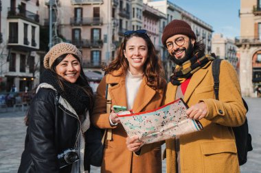 Bir grup turist, bir harita üzerinde bir şehri ziyaret edecekleri yeri izlerken kameraya bakıyor. Yolculuktaki genç insanlar gülümsüyor ve bir rehber arıyorlar.