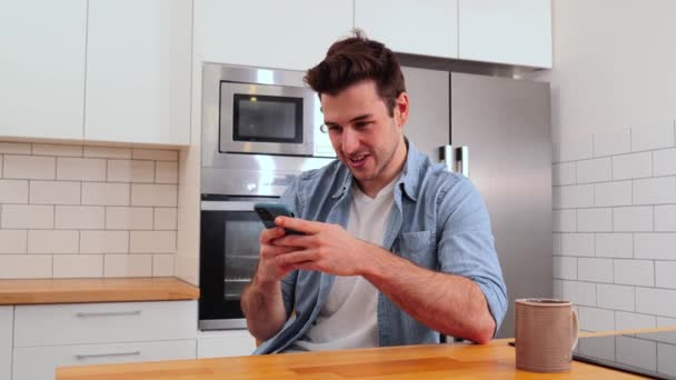 家庭のキッチンに座っている携帯電話でビデオゲームをしている若い白人男性 スマートフォンアプリを使ってSnsにメッセージを送る幸せな男 十代の男性笑顔ブラウジングとともに携帯電話 — ストック動画